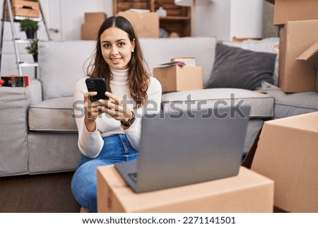 Young beautiful hispanic woman using laptop and smartphone sitting on floor at new home