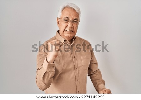 Hispanic senior man wearing glasses angry and mad raising fist frustrated and furious while shouting with anger. rage and aggressive concept. 