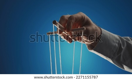 The hand of a man in a gray shirt controls a puppet using a wooden manipulator and strings on a blue background. Hand handling at puppet by pulling strings to make the character move. Close up.
