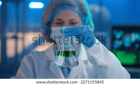 A female observes the dissolution and spread of a green substance in a flask with a clear liquid. The woman in white gown, mask, blue gloves and a bonnet observes the response of the sample. Close up.