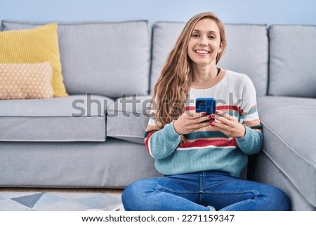 Young blonde woman using smartphone sitting on floor at home