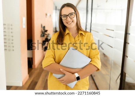 Smiling confident woman in eyeglasses looking at camera and standing in an office hallway. Portrait of confident businesswoman with a laptop, freelancer woman in coworking space