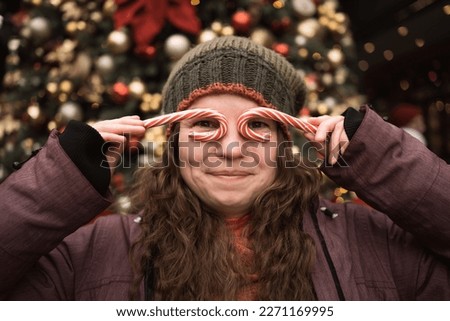 Closeup funny portrait of adult woman in hat and winter clothes makes glasses from candy canes, posing for photo at Christmas market