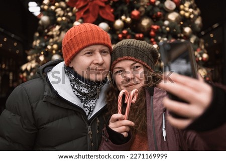Positive couple in love wearing hats and winter clothes outdoors in city street at big christmas tree background doing selfie photo by smart phone