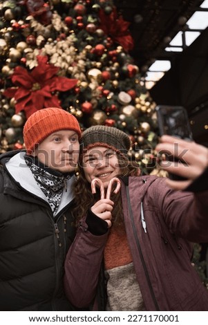 Vertical portrait of positive couple in love wearing hats and winter clothes outdoors in city street at big christmas tree background doing selfie photo by smart phone