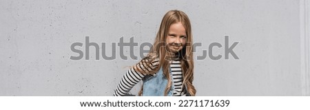 cheerful girl in denim vest striped long sleeve shirt standing near grey wall, banner