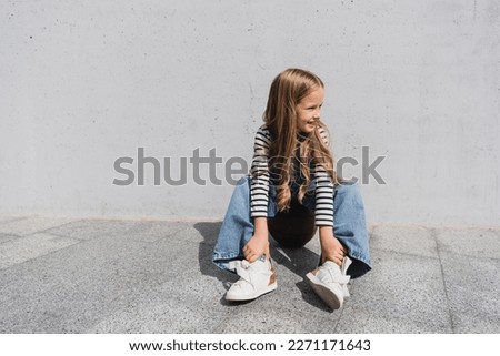 full length of smiling girl in denim vest and blue jeans sitting on basketball near wall