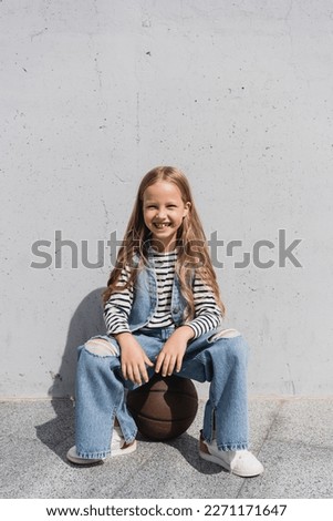 full length of cheerful girl in denim vest and blue jeans sitting on basketball near wall