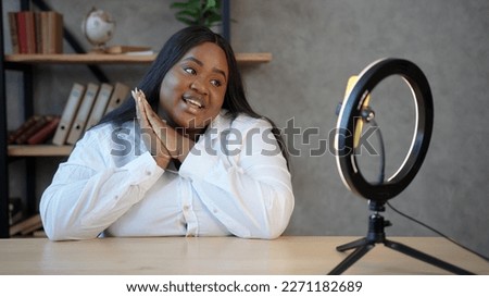 Body positive African-American beauty blogger with loose hair sits against radial light and smartphone on tripod and speaks closeup