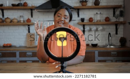 Plump blogger with dark ponytail wearing pale orange t-shirt speaks at tripod with circular lamp and golden smartphone in kitchen