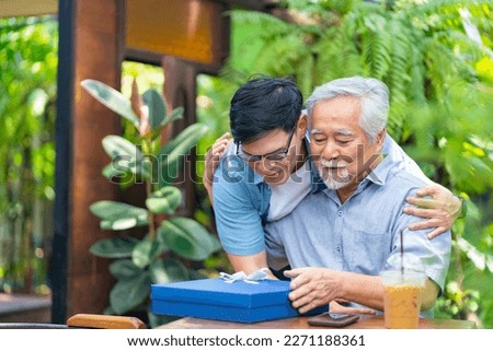 Asian man surprised elderly father with Birthday gift at outdoor cafe restaurant on summer holiday vacation. Family relationship, celebrating father's day and senior people health care concept.