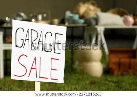 Sign Garage sale written on cardboard near tables with different stuff outdoors, closeup. Space for text
