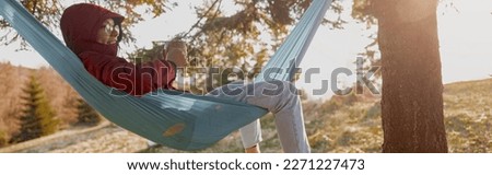 Woman in glasses and hood lying in hammock with thermos and enjoying wild nature in forest.
