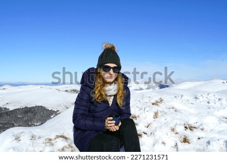 A young attractive woman with a hiking cup in her hands sits on the snow during a halt in a picturesque mountainous area