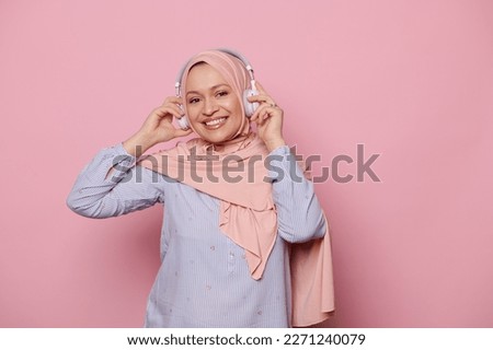 Isolated portrait on pink background of a stylish, glamour, Arab Muslim pretty woman, wearing hijab, listening to music on wireless headphones and smiling a beautiful toothy smile looking at camera
