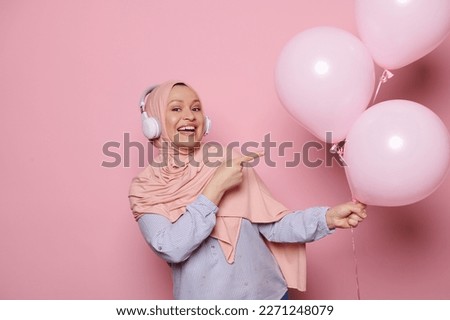 Muslim pretty woman, dressed in stylish elegant pink hijab and casual blue shirt, with headphones, pointing at a bunch of pink pastel helium balloons, smiling looking at camera on isolated background