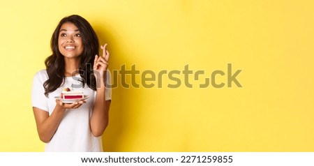 Image of cute and dreamy african-american girl, crossing fingers, holding birthday cake and looking left while making a wish, celebrating b-day over yellow background.