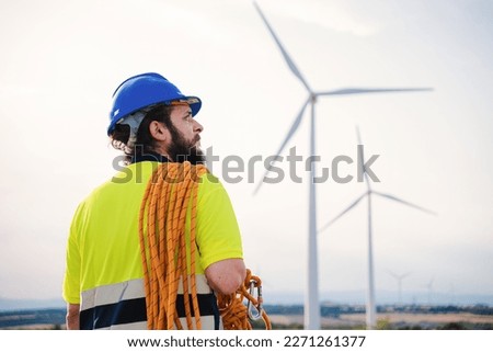 Back view of windmill turbine maintenance engineer standing wearing a hard hat nd a safety vest at wind farm with climbing equipment. High quality photo