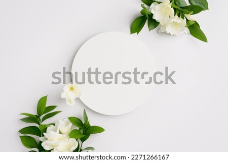 Empty round podium and white flowers with green leaves on light grey background top view. Pedestal and fresh natural green branches for cosmetic advertising. Eco product presentation mockup.