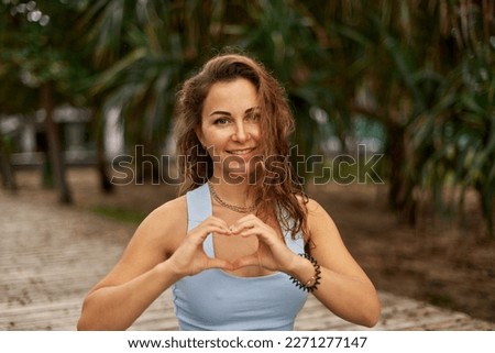 young woman in grey sport clothes making heart love hand sign, taking break during yoga exercises by green park in nature. Health care, mental health, wellbeing through meditation, sport
