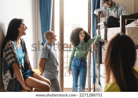 A group of diverse young adults relax in a hostel dorm room, laughing and chatting together. One individual has a shaved head, piercings, and androgynous appearance - diversity and people lifestyle Royalty-Free Stock Photo #2271288751