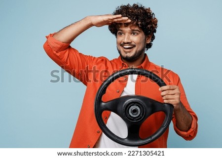 Young fun Indian man wear orange red shirt white t-shirt hold steering wheel driving car look far away distance isolated on plain pastel light blue cyan background studio portrait. Lifestyle concept Royalty-Free Stock Photo #2271305421