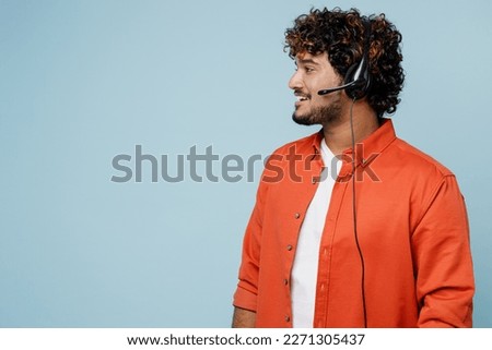 Young fun employee operator Indian business man in set microphone headset for helpline assistance wears orange red shirt white t-shirt work at call center look aside isolated on plain blue background Royalty-Free Stock Photo #2271305437