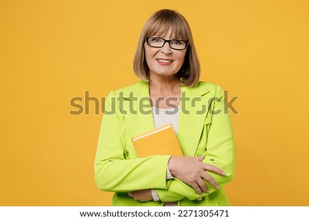 Elderly smiling happy cheerful woman 50s years old wearing green jacket white t-shirt hold in hand book novel look camera isolated on plain yellow background studio portrait. People lifestyle concept