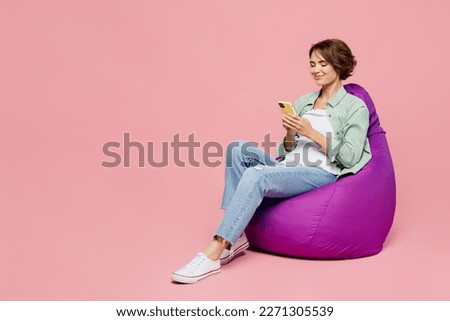 Full body young smiling happy woman 20s she wear green shirt white t-shirt sit in bag chair hold in hand use mobile cell phone isolated on plain pastel light pink background. People lifestyle concept