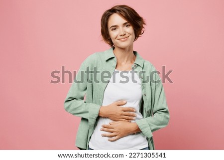 Young smiling cheerful fun happy woman 20s she wear green shirt white t-shirt holds hands on belly look camera isolated on plain pastel light pink background studio portrait. People lifestyle concept