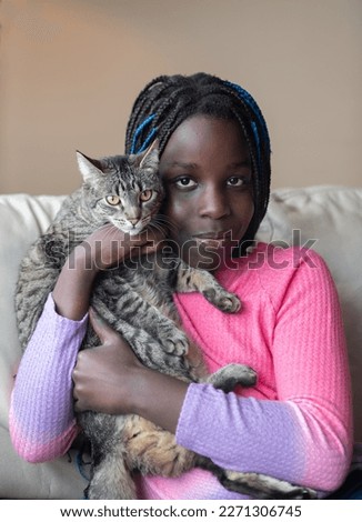 young black girl holding cat ethnic girl wearing bright pink shirt with purple sleeves holding a grey and white tabby cat indoors colored girl with long hair braids some blue in color vertical format 