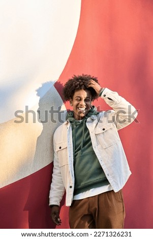 Happy cheerful African American teen guy laughing on red wall lit with sunlight. Smiling cool ethnic gen z teenager student model standing looking at camera, authentic candid shot outdoors. Vertical