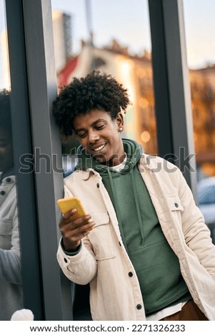 Cool smiling young African American teen guy holding mobile phone standing at glass city wall. Happy stylish hipster teen tech user using cell outdoors, looking checking apps on smartphone. Vertical