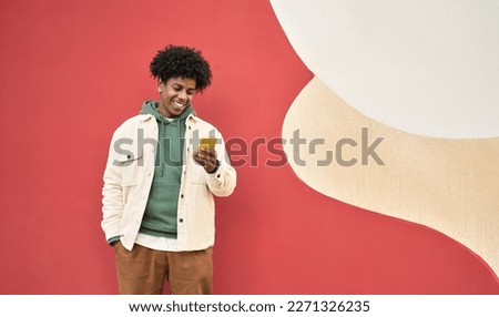 Smiling African American gen z teen standing at color wall using mobile phone. Happy cool ethnic stylish hipster guy model holding cell phone, checking apps on mobile device, looking at cellphone.