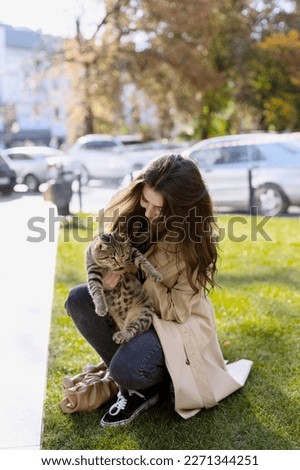 Cute, young girl holds a beautiful cat in her arms. A smiling girl on a green lawn, crouching and holding a cat, against the background of the city.