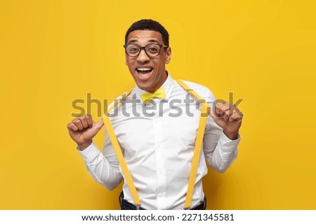 young african american guy in festive outfit with bow tie and suspenders on yellow isolated background, nerdy man in glasses smiles and poses