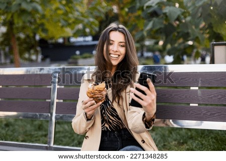 Attractive female influencer holds a cellphone and a croissant in her hands, shoots a video or takes a photo. A young woman is sitting on a bench on the street.