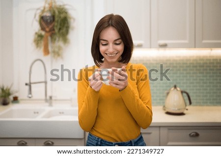 Cute cheerful pretty young brunette woman in casual outfit drinking fresh aromatic coffee at home, happy female holding tea cup in her hands and smiling, kitchen interior, copy space