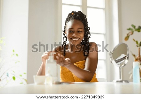Happy african american woman pouring moisturising body lotion from bottle on hand, sitting wrapped in towel, using new skincare product after bath, free space Royalty-Free Stock Photo #2271349839