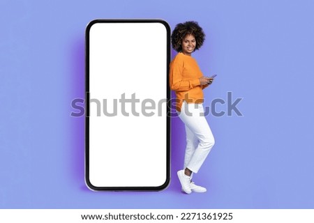 Positive cheerful stylish beautiful young african american lady leaning on huge phone with white blank screen, using smartphone and smiling at camera, purple background, enjoying newest app, mockup
