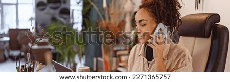 Smiling female florist working on laptop in floral studio, using phone for check customers order