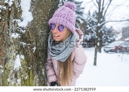 blond-haired woman with winter clothes and sunglasses standing next to a tree in the snow, medium shot. High quality photo