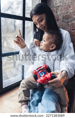 Caucasian young woman sitting on the window-sill with his little son and showing something through the window. High quality photo