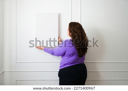 Attractive fat woman seen from behind putting a picture frame or mockup on the wall while decorating her home