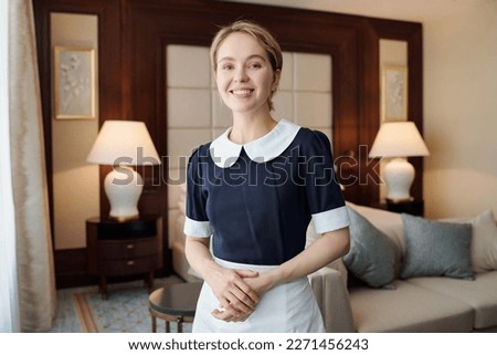Young successful chambermaid in uniform standing in front of camera against couch and two lamps in room of luxurious hotel Royalty-Free Stock Photo #2271456243