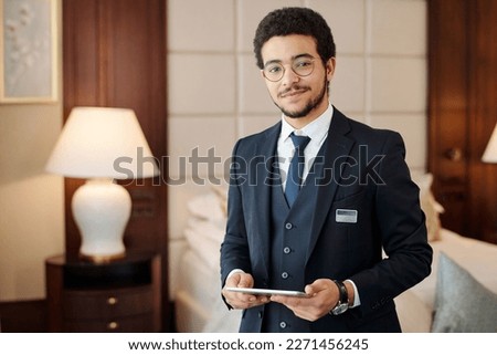 Young confident head manager of luxurious hotel looking at camera while standing against lamp and comfortable double bed in room Royalty-Free Stock Photo #2271456245