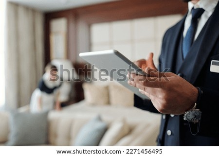Close-up of young male manager of hotel using digital tablet while looking through list of booked guests with chambermaid on background