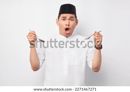 Surprised young Asian Muslim man in Arabic clothes holding spoon and fork with open mouth isolated on white background. People religious Islamic lifestyle concept
