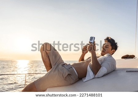 Asian young man tourist using smartphone taking photo during yachting. Attactive male feel happy and relax while sitting alone on deck of yacht, enjoy time when catamaran boat sailing during summer.