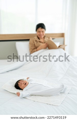 Newborn baby lie on bed with relax and happiness while her Asian mother sit and read book in bedroom in background. 